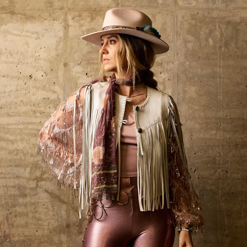 A woman modeling a fringed leather vest, sheer patterned sleeves, and metallic pink pants, accessorized with a wide-brimmed hat and scarf.