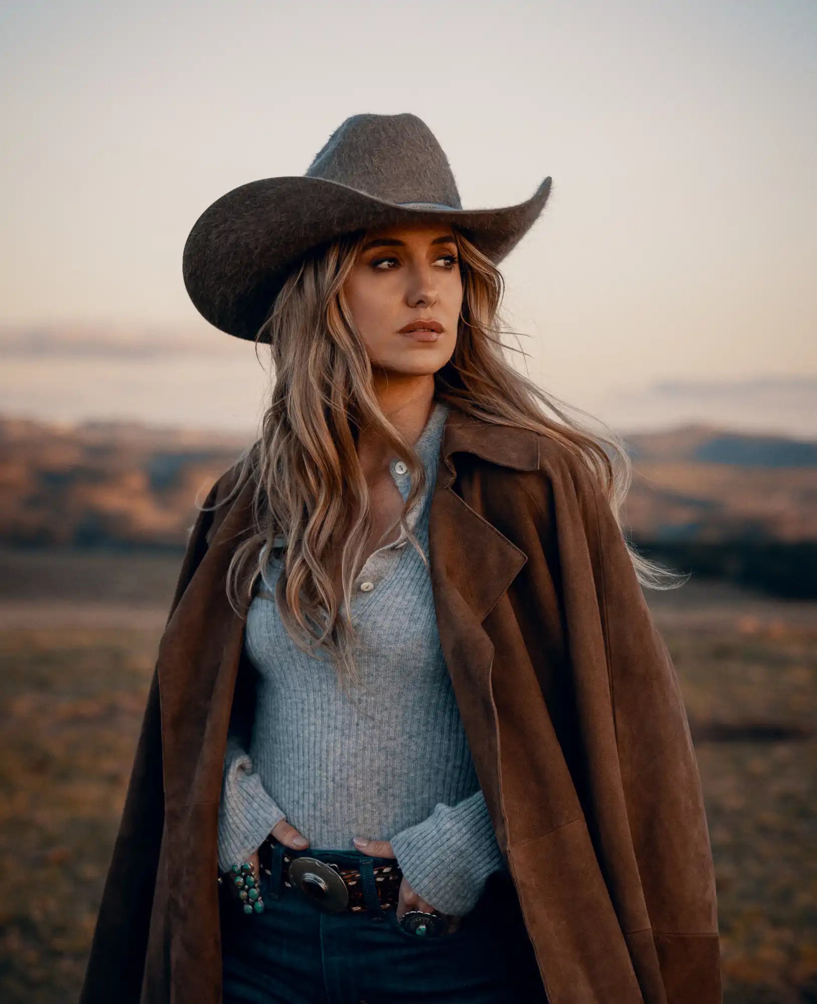 A woman wearing a brown cowboy hat, a light grey Henley sweater, and a brown coat.