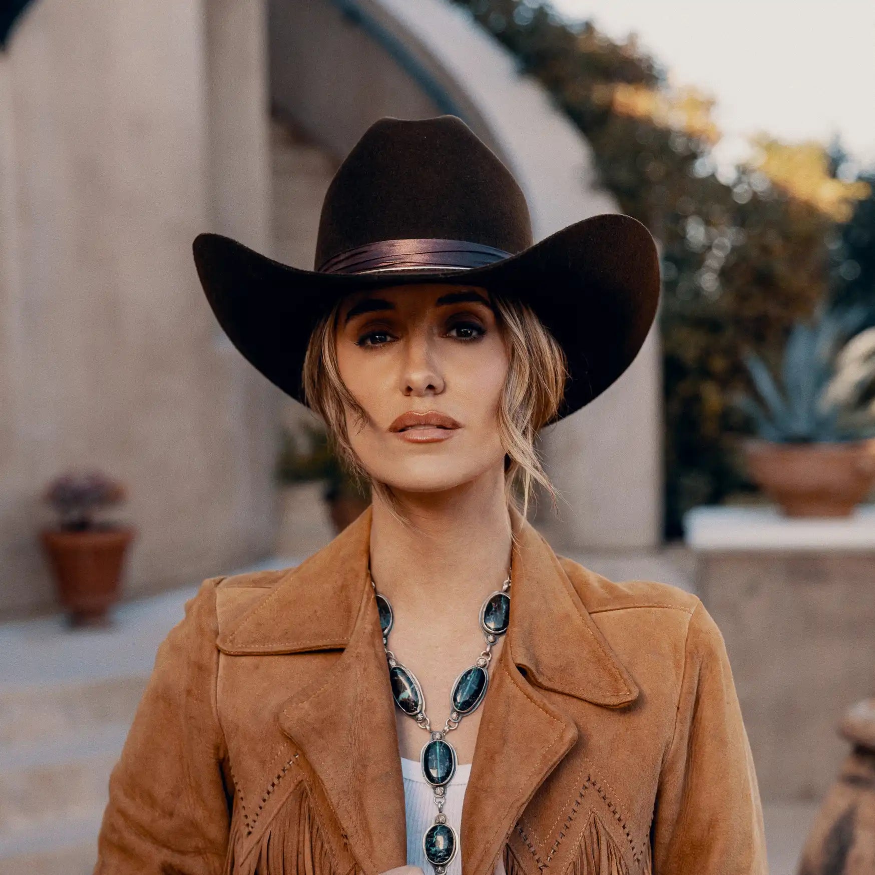 A woman in a brown cowboy hat, a tan fringed jacket, and a long silver and black stone necklace.